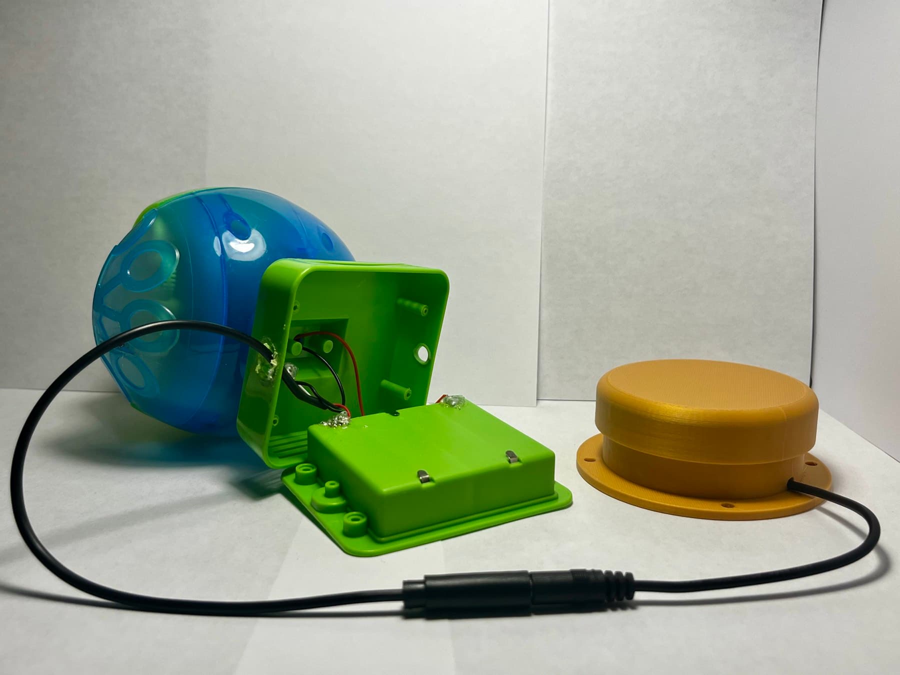 The rear of a bubble machine with its cover off, showing wiring routed through a 3D-printed junction box to a round yellow activation switch.