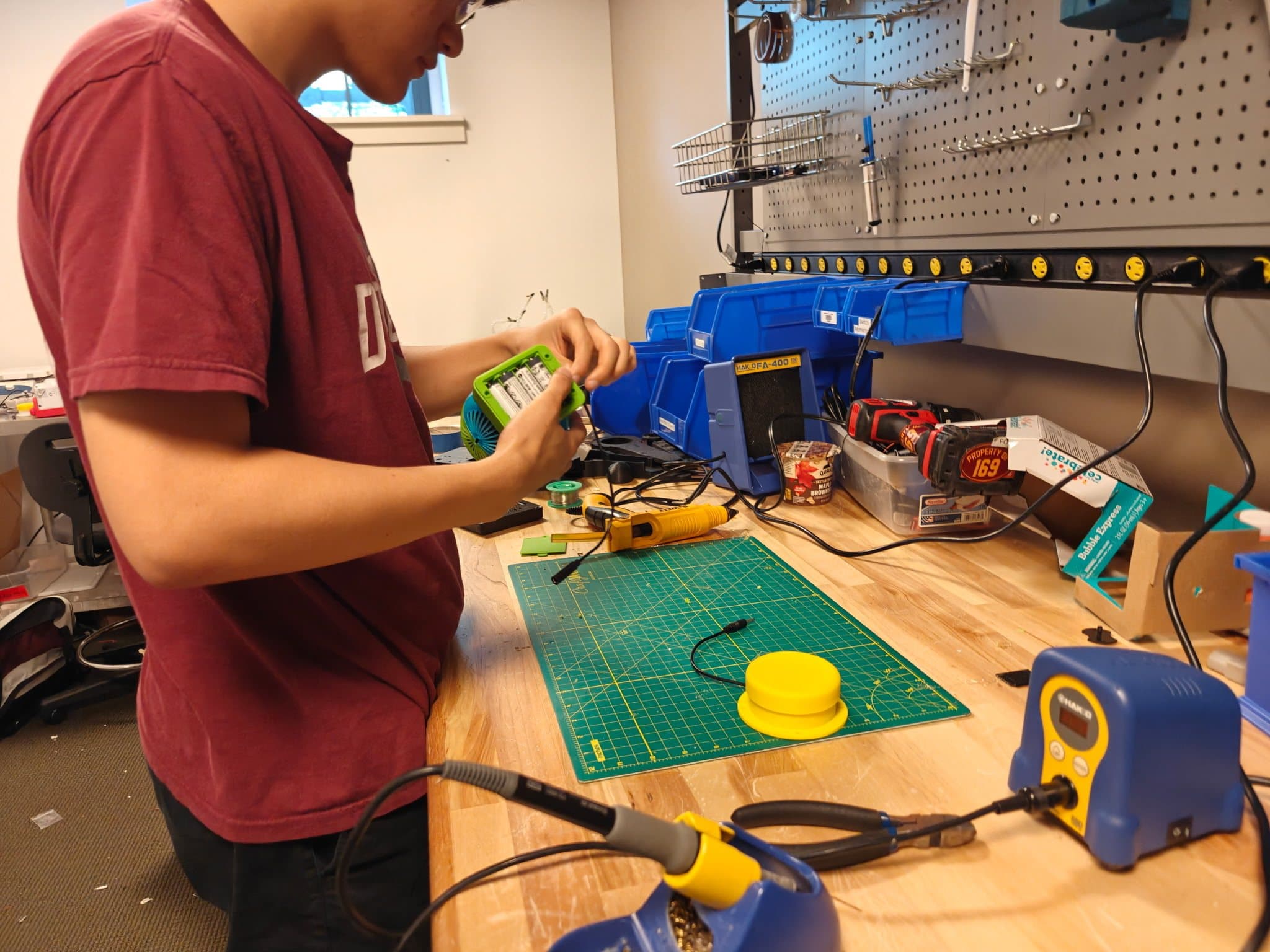 A student at a makerspace workbench wiring the battery compartment of a green bubble machine, with a yellow assistive switch on the cutting mat beside them.
