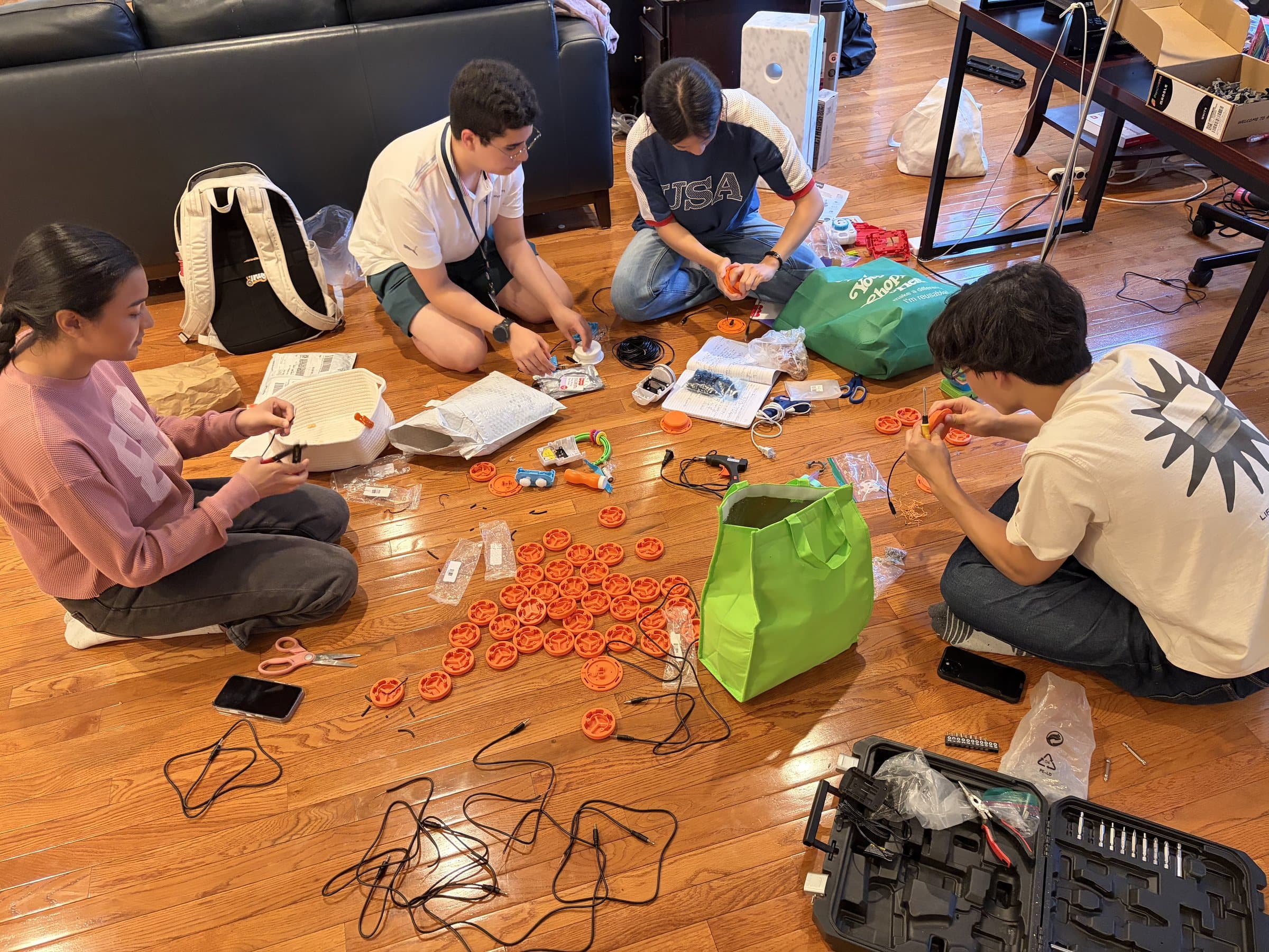 Four team members sitting on a living room floor assembling orange switch buttons, surrounded by cables, tools, and open notebooks.