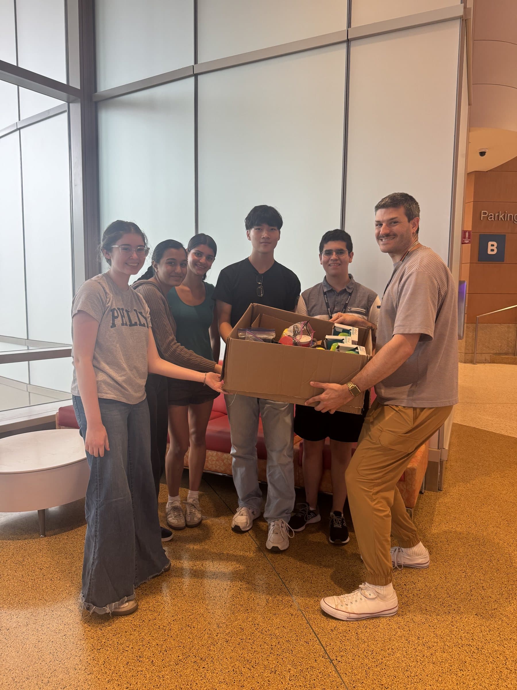 Five Adapt Project students and Daniel Fichter holding a cardboard box packed with adapted toys and printed devices in a hospital lobby.