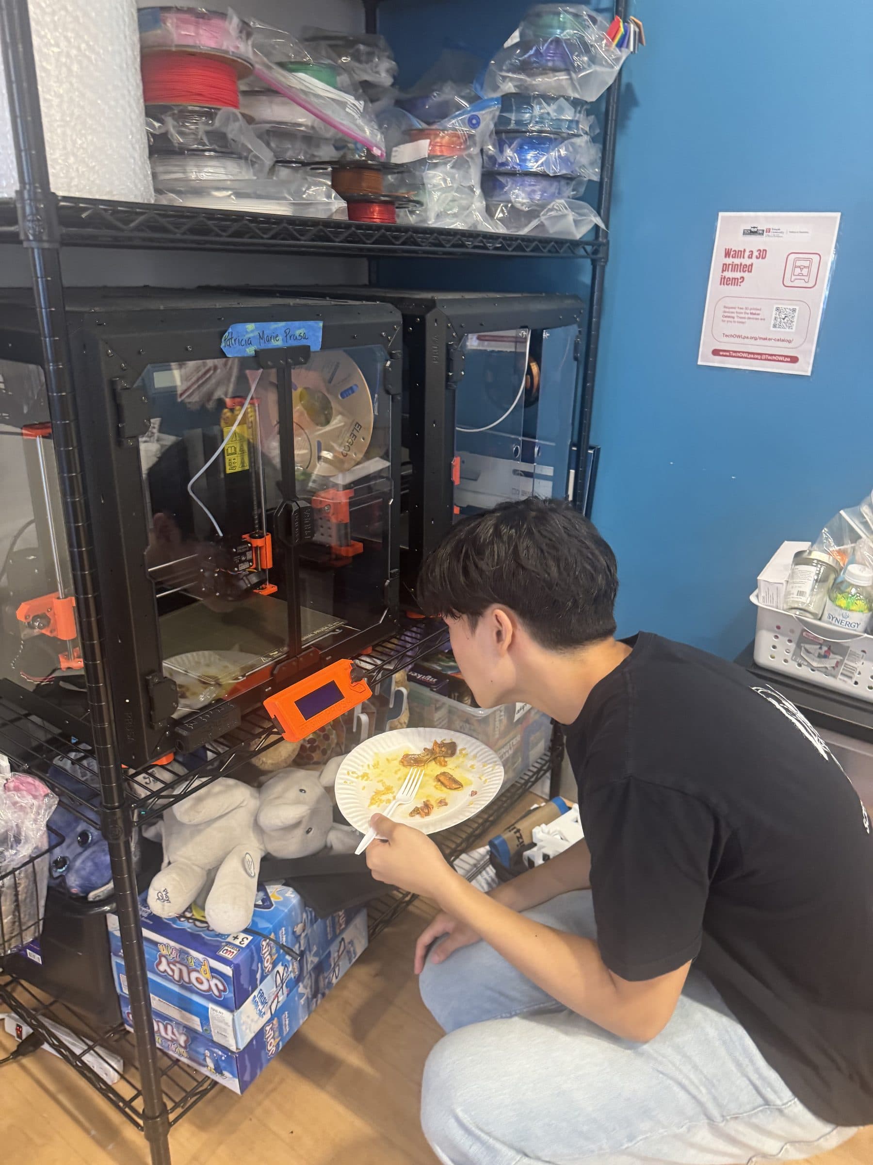 An Adapt Project member crouched beside a bank of 3D printers at TechOWL, watching a print run, with bagged filament spools stacked on the shelf above.