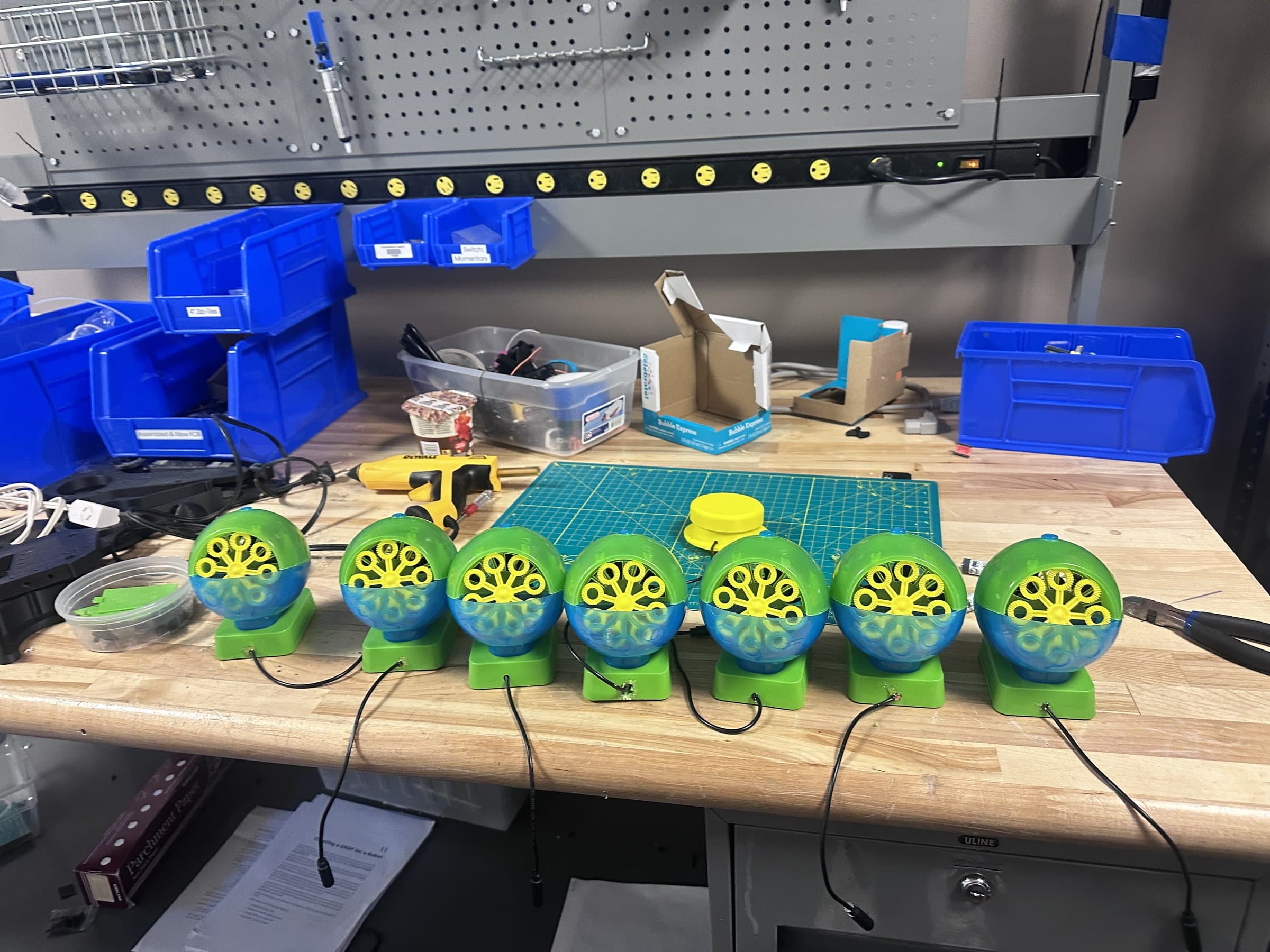 Seven switch-adapted bubble machines lined up on a workbench, each with a 3.5mm jack cable soldered in.