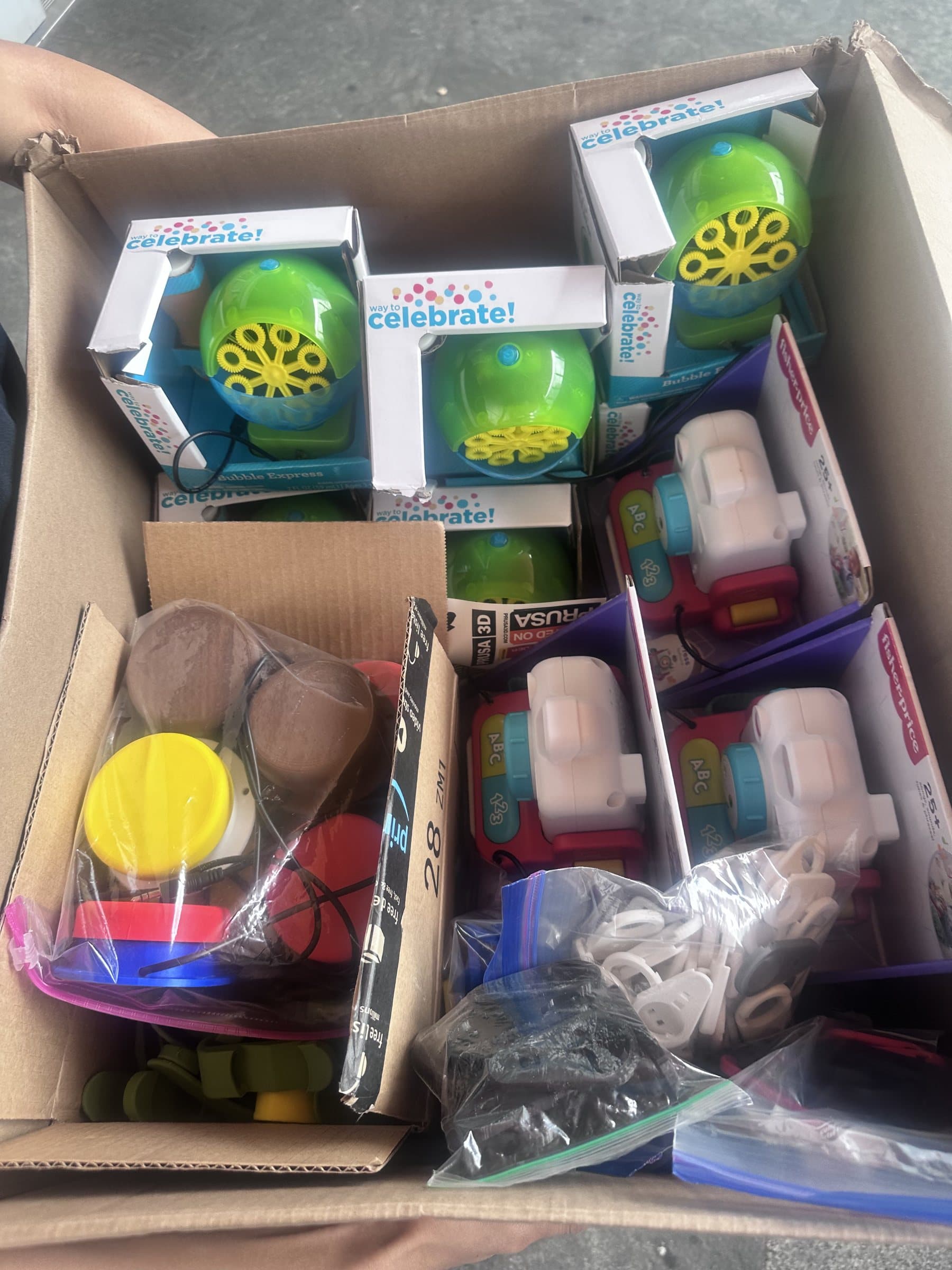 A cardboard box packed with boxed bubble machines, Fisher-Price camera toys, and bags of 3D-printed switch buttons and zipper pulls.